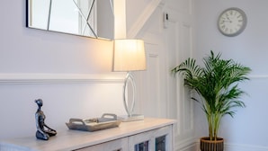 A hallway with a light-colored console table, a wall-mounted clock, a mirror, a lamp, a potted plant, and a decorative sculpture. - 1 Bed in Colwyn Bay (oc-78154) (Hen Golwyn)