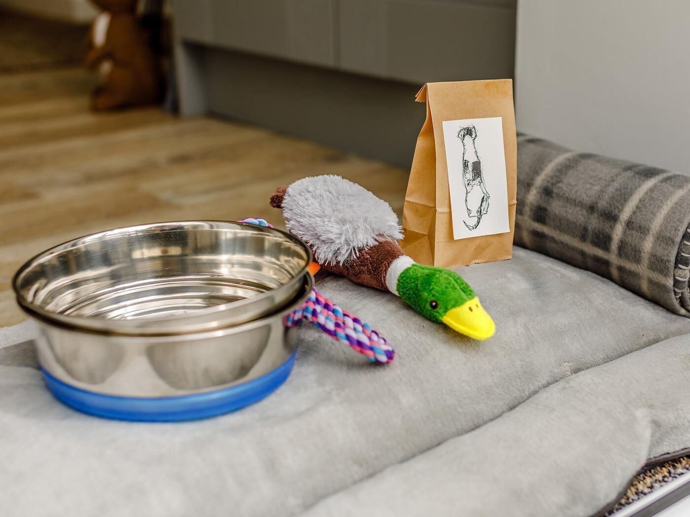 Une gamelle en métal pour chien, un jouet en forme de canard en peluche et un sac en papier kraft avec un dessin dessus.