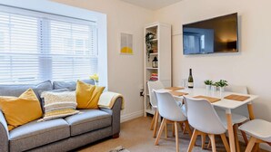 A modern living room with a grey sofa, white chairs, a dining table, a bookshelf, and a flat-screen TV. - 3 Bed in Ventnor (oc-80547) (Ventnor)