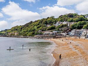 Ein Küstenstrand mit Häusern an einem Hügel, bewölkter Himmel und ruhigem Wasser.