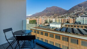 Ein Balkon mit Tisch und Stühlen, Blick auf eine Stadtlandschaft mit Gebäuden und Bergen, sowie Solaranlagen auf dem Dach.