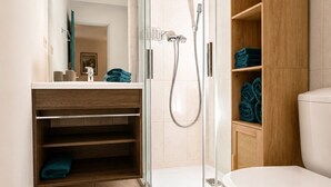 Un baño moderno con cabina de ducha de vidrio, tocador de madera, inodoro blanco y alfombra color turquesa.
