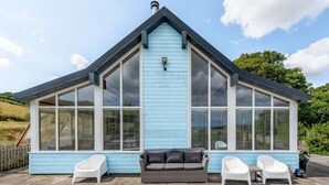 Una casa azul con un amplio ventanal frontal, techo negro y patio de piedra con mobiliario exterior.