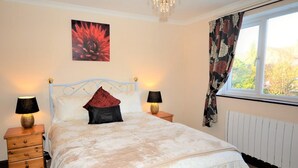 Een slaapkamer met een bed, twee nachtkastjes, een lampje, een kroonluchter, een raam met gordijnen en een aan de muur bevestigd schilderij.