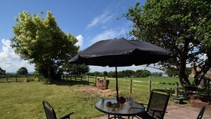 Een terras buiten met een tafel, stoelen, een zwarte parasol en uitzicht op een grasveld en bomen.