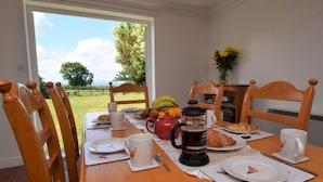 Een eettafel met stoelen, een koffiepot, borden, een croissant en uitzicht op een groen landschap door een raam.