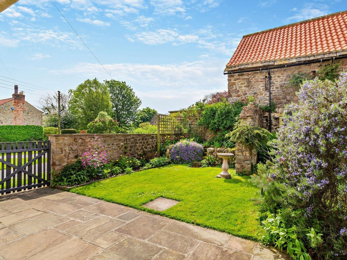 A stone building with a tiled roof, a stone wall, and a well-maintained garden with a lawn, a birdbath, and flowering plants.