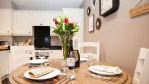 A dining table set with plates, cutlery, and a bottle of wine, surrounded by white chairs in a kitchen with cabinets and a clock on the wall. - 2 Bed in Stokesley (oc-g0072) (Great Busby)