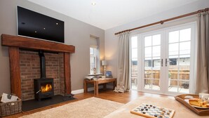 A living room with a fireplace, a television mounted on the wall, a wooden table, and a large window with curtains. - 2 Bed in Stokesley (oc-g0072) (Great Busby)