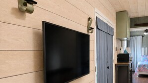 Ett modernt rum med en väggmonterad TV, väggpaneler i trä, en dörr med gardiner, ett litet bord med en lampa och en säng med ett blommigt överkast.