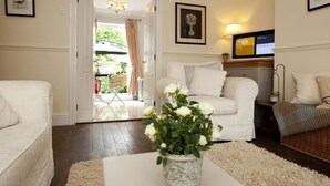 Ett vardagsrum med en vit soffa, en vit fåtölj, ett soffbord med en vas med blommor, en tv och en dörr som leder ut till ett utomhusområde.