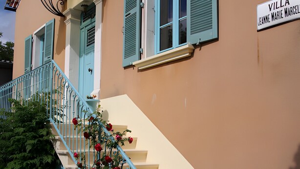 A building with a sign reading 'VILLA JEANNE MARIE MARCELLE,' a staircase with blue railings, and red flowers.