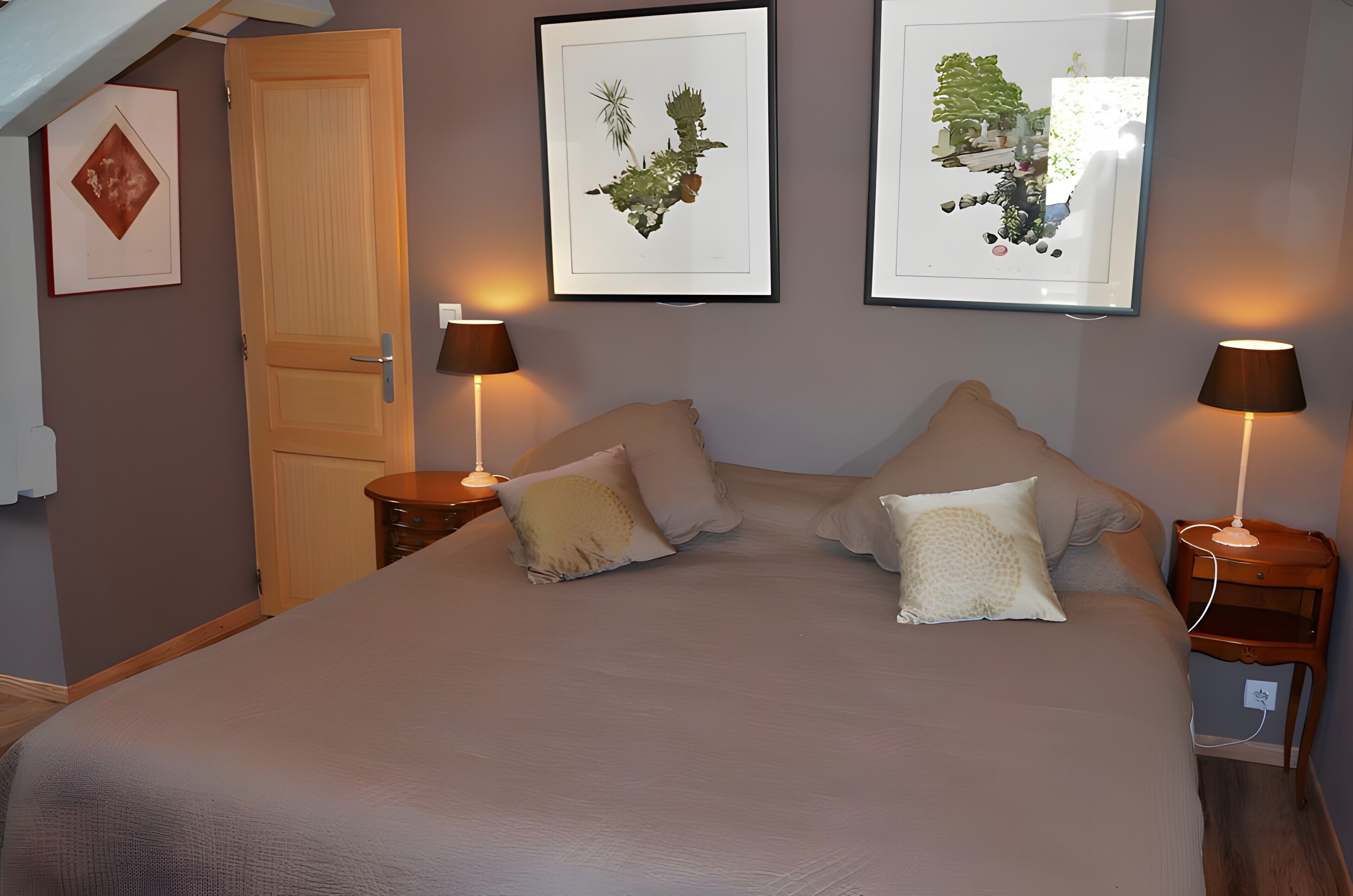 A bedroom with a bed, two bedside tables with lamps, a door, and two framed botanical prints on the wall.