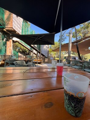 An outdoor seating area with wooden tables and chairs, umbrellas providing shade, and a view of the surrounding trees and buildings. - The Seagulls of the Coast (Saint-Jean-de-Monts)