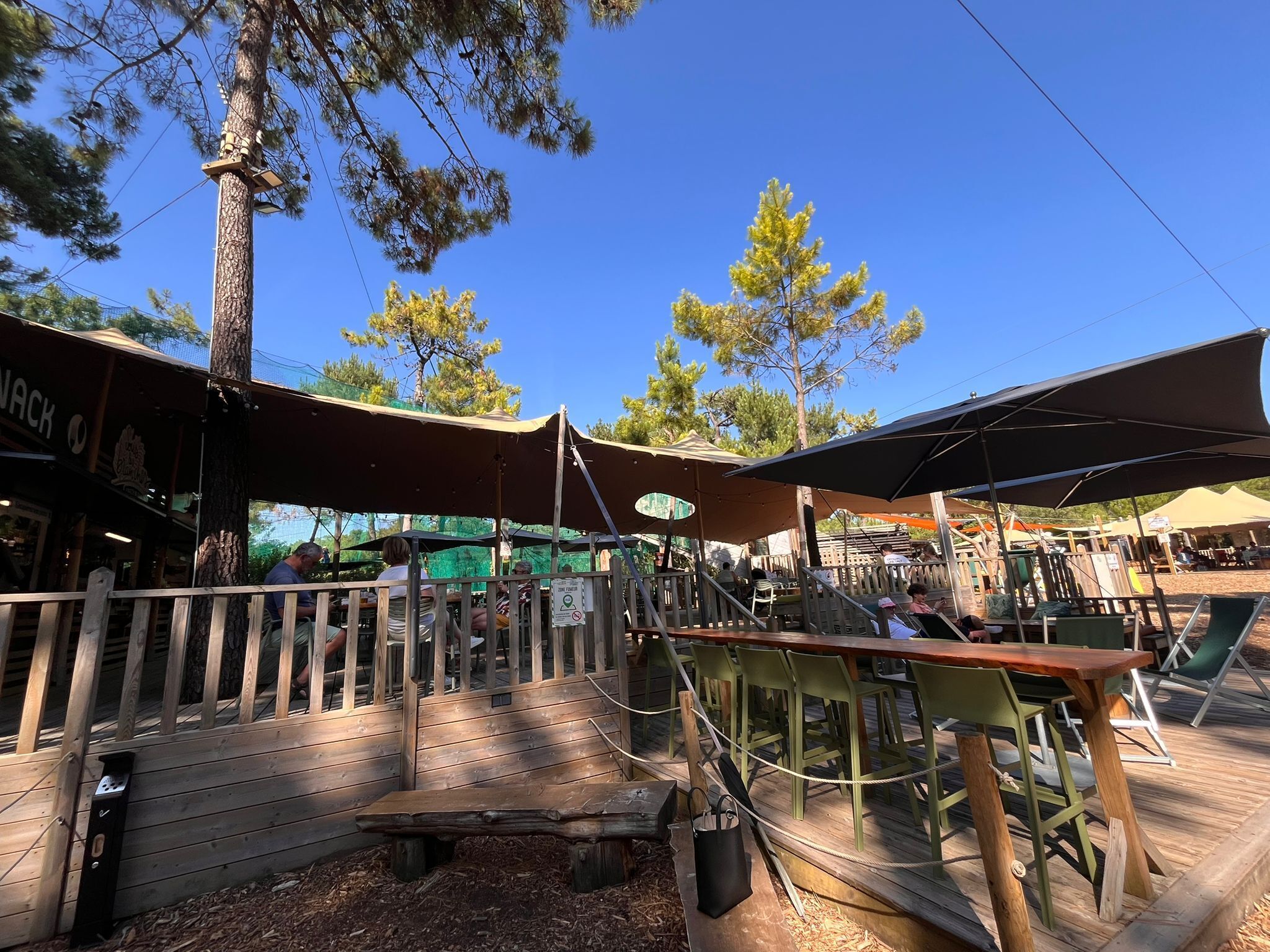 An outdoor seating area with wooden tables and chairs, surrounded by trees and covered by canopies.