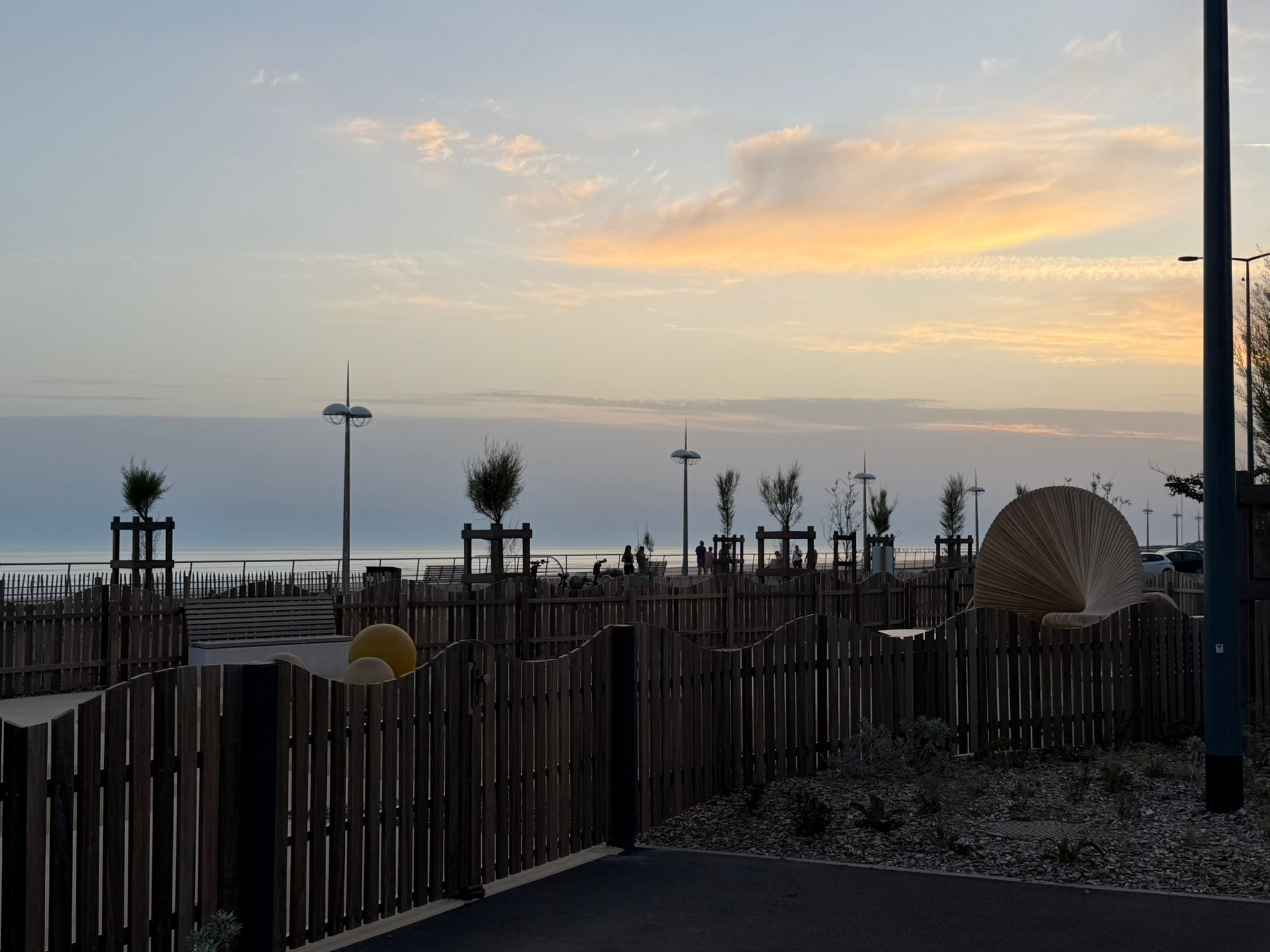 A wooden fence with a gate, a paved walkway, and a view of the sea.