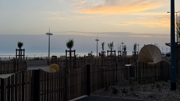 Una valla de madera con un portón, un sendero pavimentado y vista al mar.