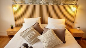 Una cama bien hecha con almohadas, una manta y toallas. Hay dos mesitas de noche con lámparas y pequeñas plantas.