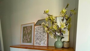 Un gabinete blanco con repisa superior de madera que sostiene obras de arte enmarcadas y un florero con flores.