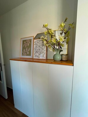 Een witte kast met een houten bovenblad waarop ingelijste kunstwerken en een vaas met bloemen staan.