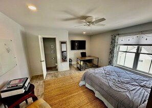 Ein Schlafzimmer mit Bett, Schreibtisch, Stuhl, Deckenventilator, einem Fenster mit Vorhängen und einer Tür zu einem anderen Raum.