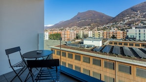 A balcony with a table and chairs, a view of a cityscape with buildings and mountains, and solar panels on the roof. - {Suite 410} Gym, Business Room, Modern, Relax (Lugano)