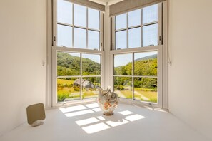 A room with a large window overlooking a green landscape, a white vase, and a stone on the floor.