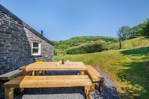 A stone building with a wooden table and benches, a window, and a view of green hills.