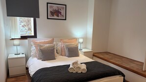 A bedroom with a bed, two bedside tables with lamps, a window with a view, a framed picture on the wall, and a wooden bench.