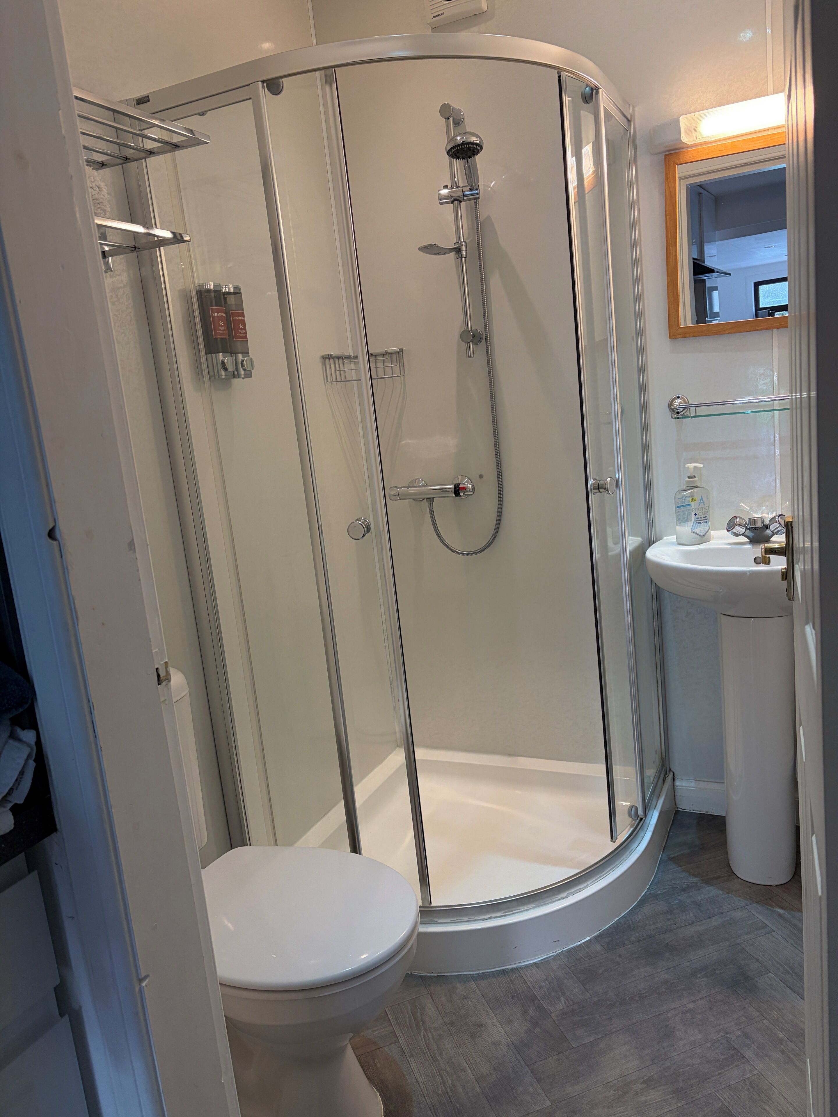 Et moderne badeværelse med en glasvæg omkring brusenichen, et hvidt toilet og en vask med et spejl over.