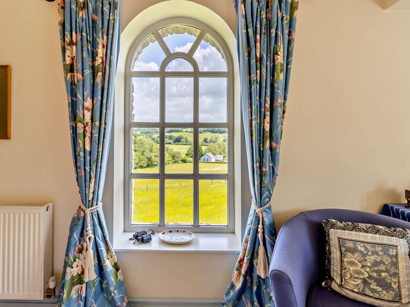 A room with a large arched window, curtains, a radiator, a plate, and a cushion.