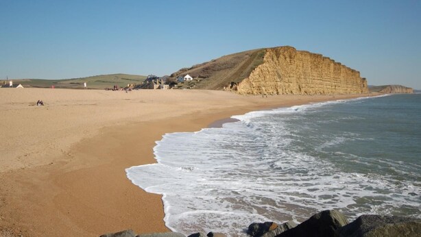 Ein Strand mit einer markanten Klippe, einem Sandstrand und einigen Gebäuden im Hintergrund.