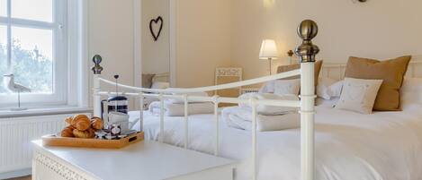 A hotel room with a white bed, a bedside table with a tray of croissants, a window with a view, and a wall-mounted heart decoration.