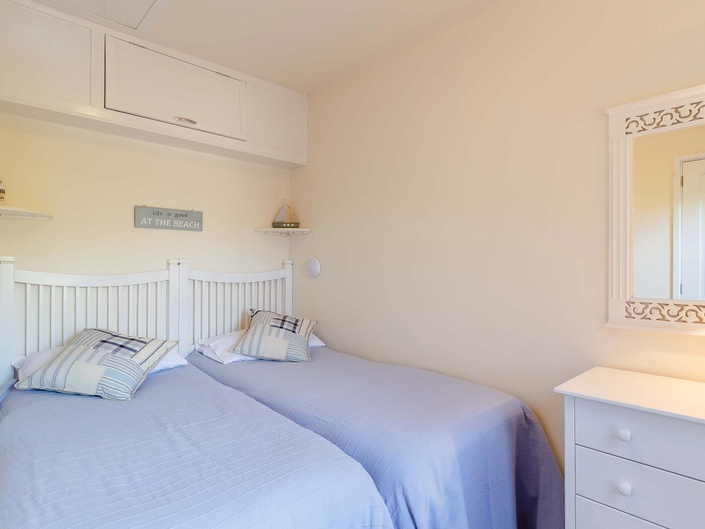 A bedroom with two beds, a white dresser, a mirror, a shelf with a decorative item, and a sign on the wall.