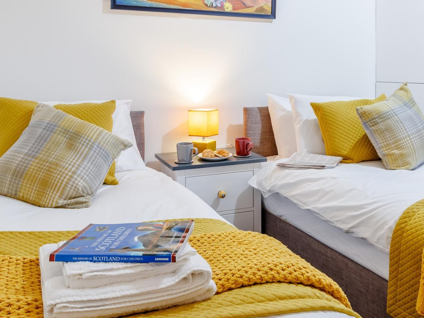 A hotel room with two beds, a nightstand with a lamp, a red mug, and a book titled 'The History of Scotland for Children'.