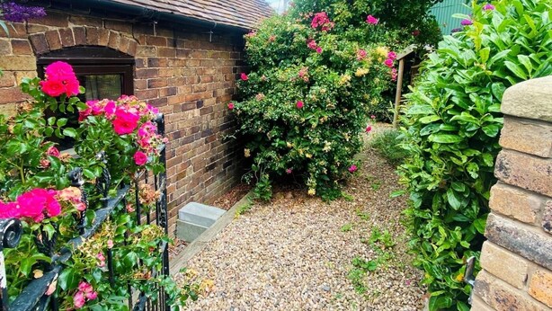 A brick building with a sloped roof, surrounded by lush greenery and flowering plants. - 1 Bed in Leighton (oc-80726) (Leighton)