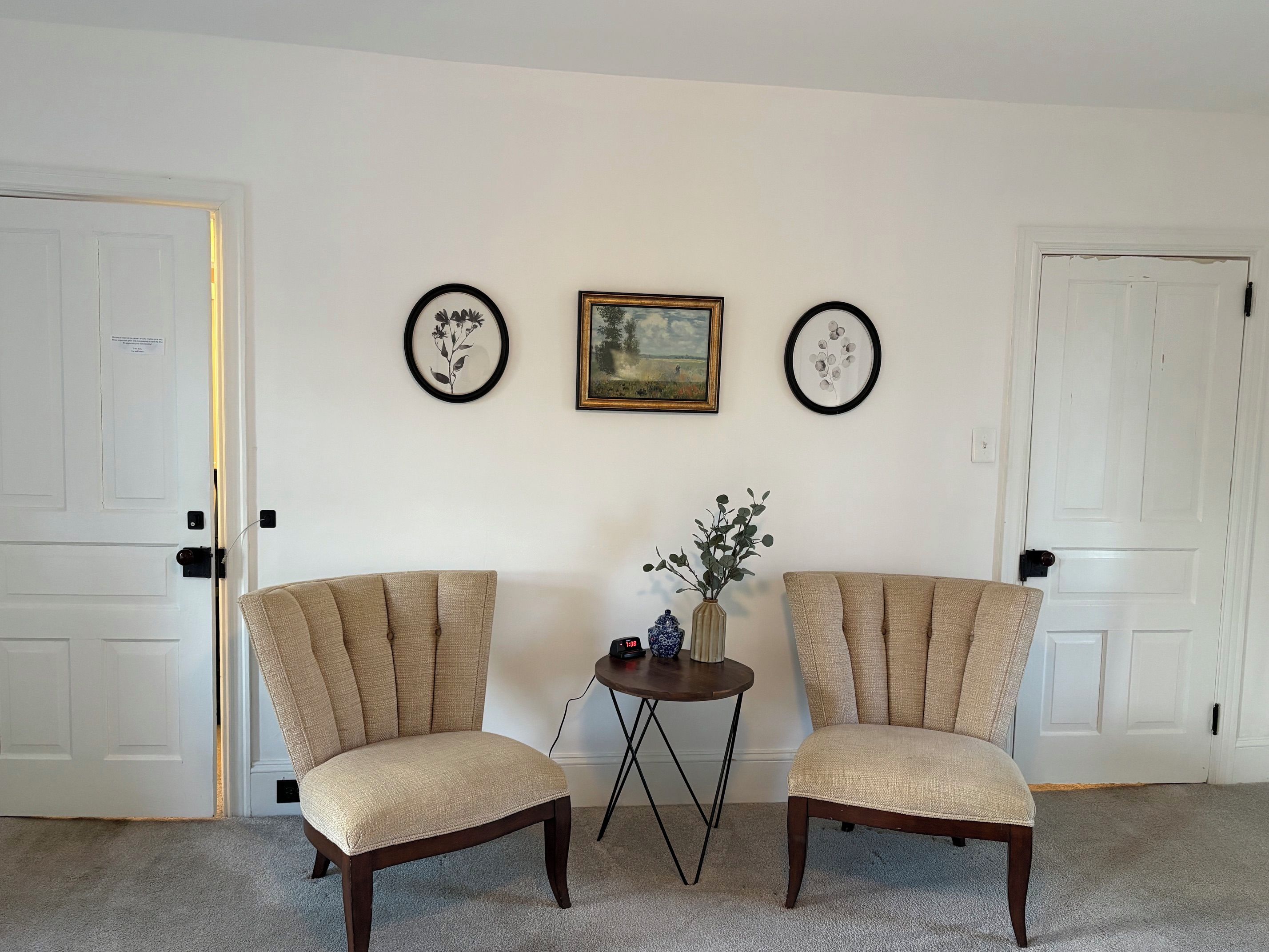 A room with two beige armchairs, a small round table with a vase and a clock, and two framed pictures on the wall.