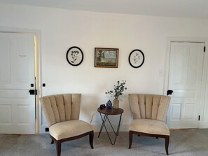 A room with two beige armchairs, a small round table with a vase and a clock, and two framed pictures on the wall.