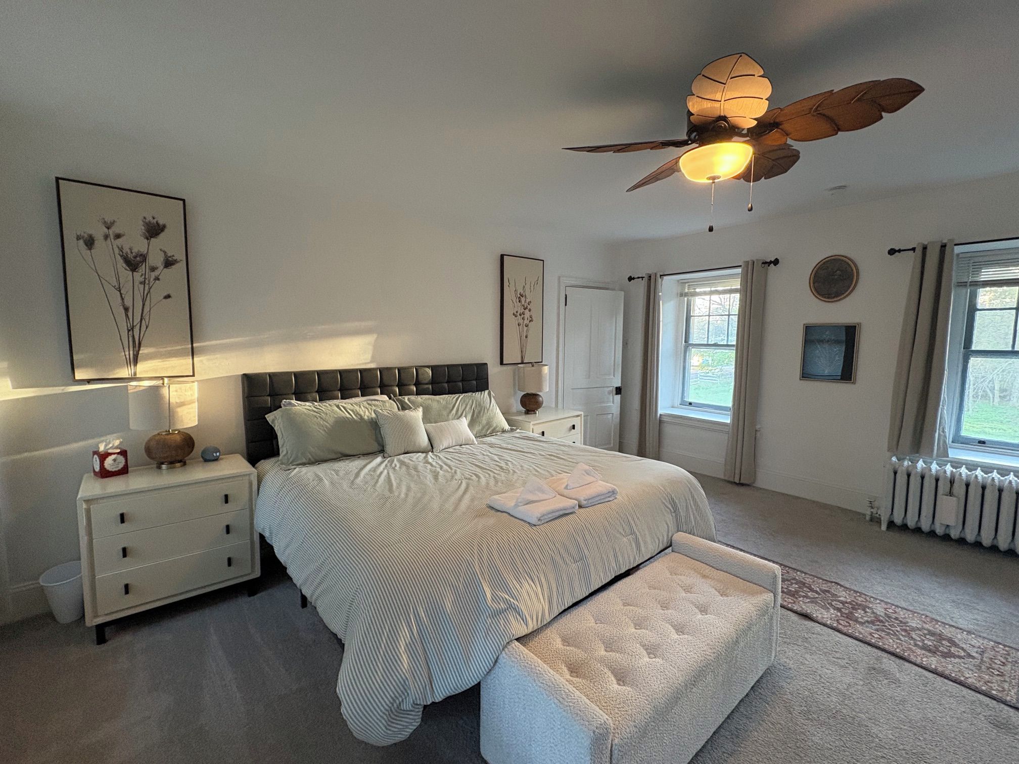 A bedroom with a large bed, bedside tables, a nightstand, a ceiling fan, and a window with curtains.