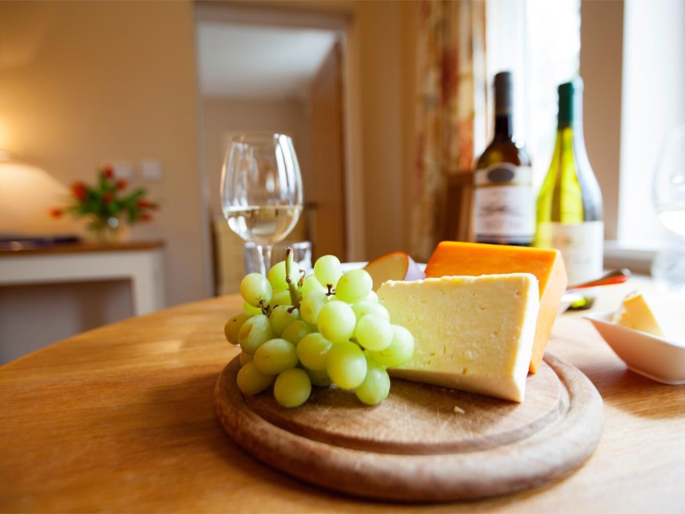 A wooden table with a cheese and grapes, a wine glass, and bottles in the background.