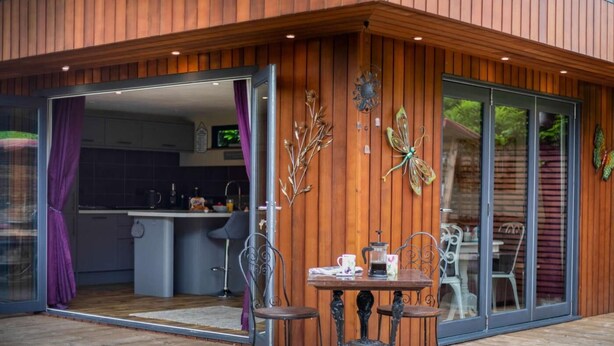 Una casa moderna con fachada de madera, puerta de cristal y una zona de comedor con sillas metálicas. Se ve una cocina a través de la puerta.