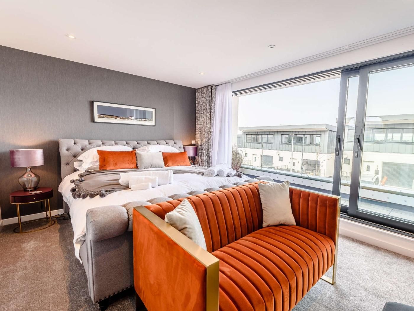 A modern hotel room with a large bed, a sofa, a view of the city through large windows, and a framed picture on the wall.