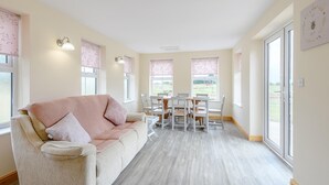 Ein gemütliches Wohnzimmer mit einem beigen Sofa, einem Esstisch mit Stühlen und großen Fenstern mit floralen Rollos.