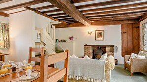 Ein gemütliches Wohnzimmer mit freiliegenden Holzbalken, einem Kamin, einer Essecke mit gedecktem Tisch für das Frühstück und einem bequemen Sofa.