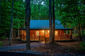A cabin with a wooden deck, large windows, and a metal roof, surrounded by tall trees. - Cedar Cabin Stay (Sevierville)
