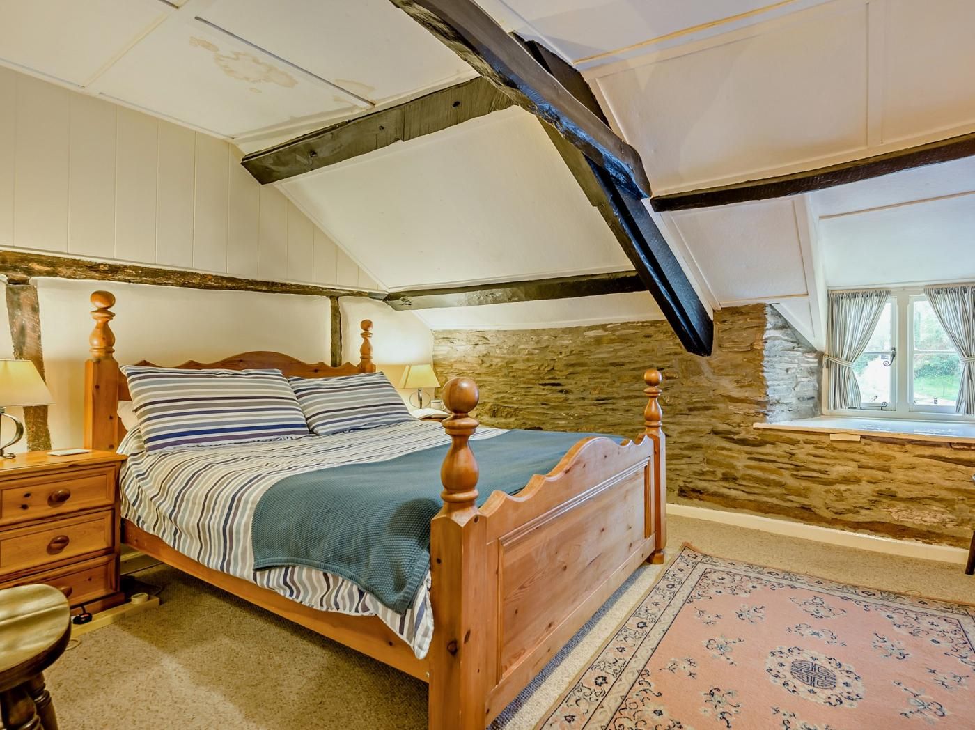 Un dormitorio con techo inclinado, cama de madera con cabecera, mesita de noche, lámpara, pared de piedra y ventana con cortinas.