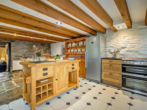 A kitchen with wooden cabinets, a refrigerator, an oven, and a stone wall. - 3 Bed in Boscastle (oc-75145) (Boscastle)