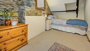 Ein Schlafzimmer mit Stehwänden, einem hölzernen Schminktisch, einem Nachttisch mit Lampe, einem Bett mit gestreifter Bettwäsche und einem kleinen Teppich.