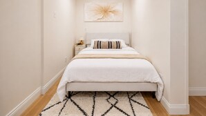Habitación de hotel estrecha con cama, mesita de noche, un cuadro colgado en la pared y una alfombra estampada.