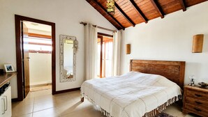 A bedroom with a wooden bed, a nightstand, a mirror, a door leading to a bathroom, and a ceiling with wooden beams. - Villa Serena | Vacation Home for Families and Groups (Armação dos Búzios)